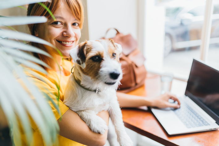 Mulher sorrindo enquanto segura um cachorrinho (filhote de porte pequeno) no colo, em um ambiente de trabalho com notebook sobre a mesa e outras pessoas ao fundo.